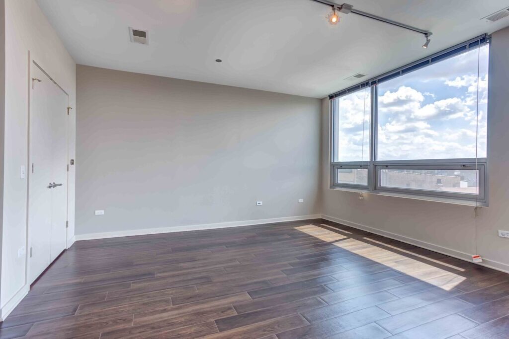 Empty room with large windows, wooden flooring, light-colored walls, and a ceiling with a single track light. Bright daylight streams through the windows, illuminating the space.