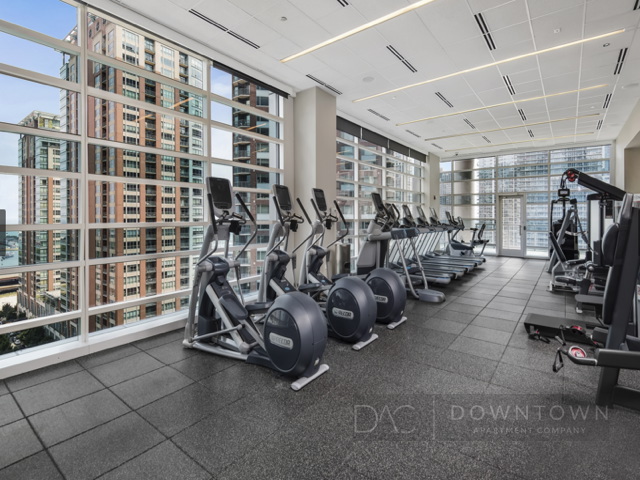 A modern gym with large windows overlooking a cityscape, featuring elliptical machines, treadmills, and other fitness equipment on a tiled floor.