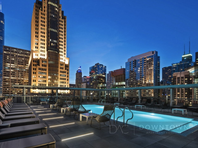 Rooftop pool area at night surrounded by city skyscrapers with lounge chairs lining the poolside.