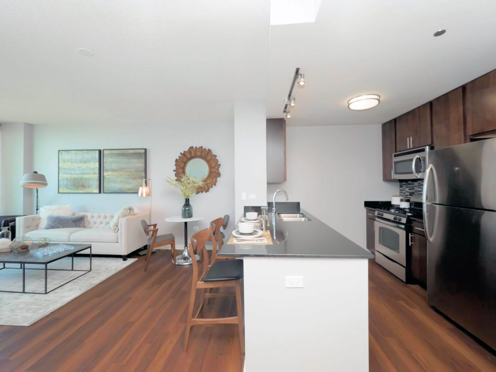 Modern open-plan kitchen and living area with wooden flooring, dark cabinets, stainless steel appliances, and a breakfast bar with stools; art pieces and decor accentuate the space.