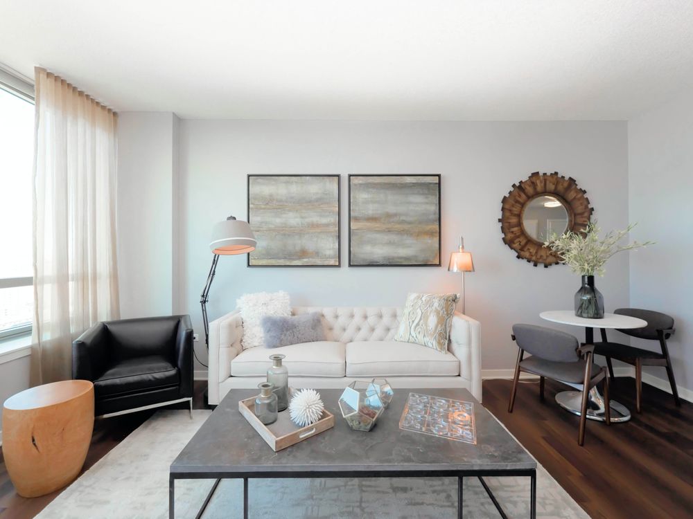 Modern living room with a white sofa, black chair, and round table with two chairs. Two abstract paintings hang on the wall. A large window with sheer curtains lets in natural light.