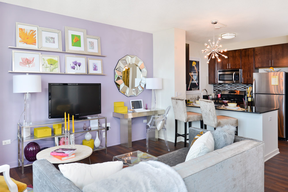 Modern living room with a gray sofa, glass coffee table, and wall-mounted TV. Purple accent wall with floral artwork. Open kitchen with dark cabinets and a small dining area with beige chairs.