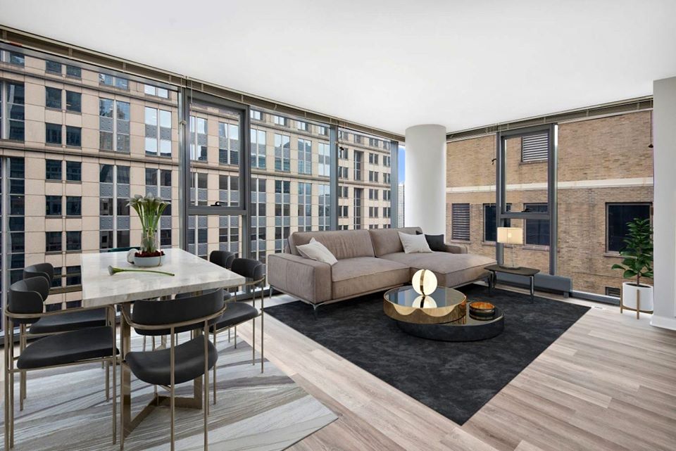 Modern open-plan apartment with large windows. Features a dining table with chairs, a gray sofa, a coffee table, and a small plant. Overlooks tall city buildings. Neutral color palette.