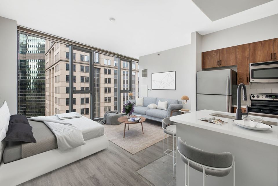 Modern studio apartment with large windows, a bed, sofa, coffee table, and kitchen area. High-rise buildings visible outside. Neutral tones and sleek finishes dominate the decor.