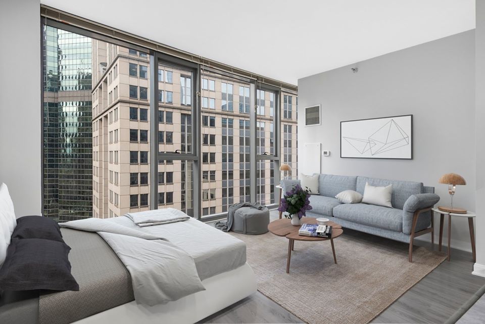Modern studio apartment with a bed, sofa, and coffee table. Large windows offer a view of adjacent skyscrapers. Neutral color scheme with minimalist decor and a wall art piece.