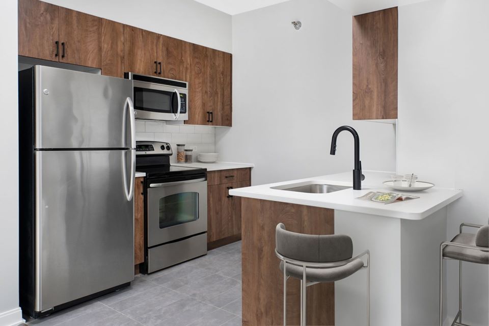 Modern kitchen with stainless steel appliances, wooden cabinets, and a white island featuring a sink and two bar stools. Gray tiled floor and minimalistic decor.