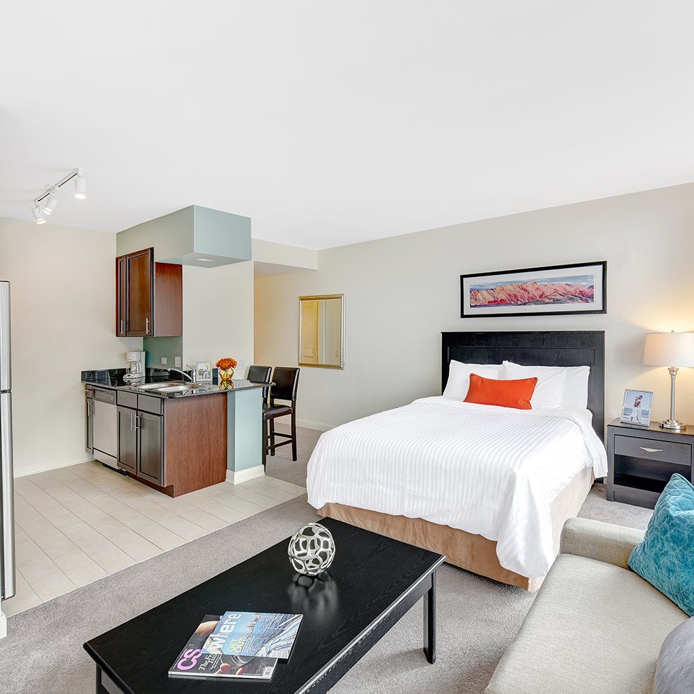 Modern studio apartment with bed, kitchen area, and seating. Includes a black coffee table with magazines and decor, cozy sofa, bedside lamp, and framed wall art. Neutral tones with vivid accents.