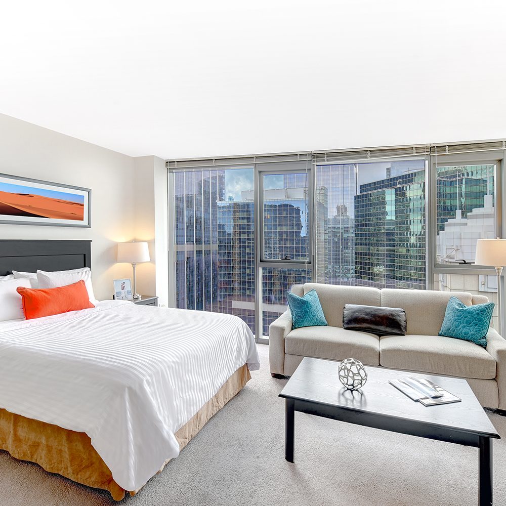 Bright modern hotel room with a large bed, colorful pillows, a sofa, and a coffee table. Floor-to-ceiling windows offer a cityscape view of tall buildings.