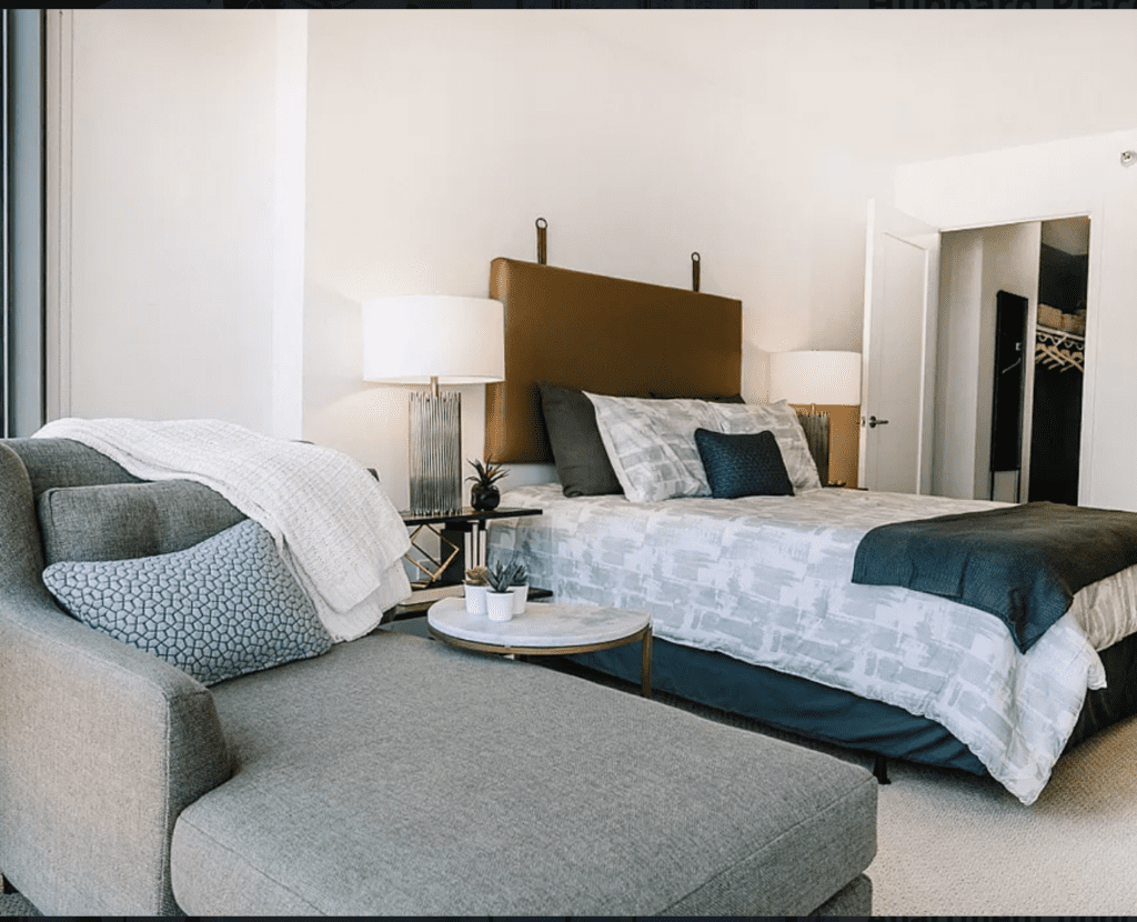 A modern bedroom featuring a large bed with patterned bedding, a cushioned headboard, and bedside lamps. A gray sofa with pillows and a throw blanket is in the foreground, along with a small table.