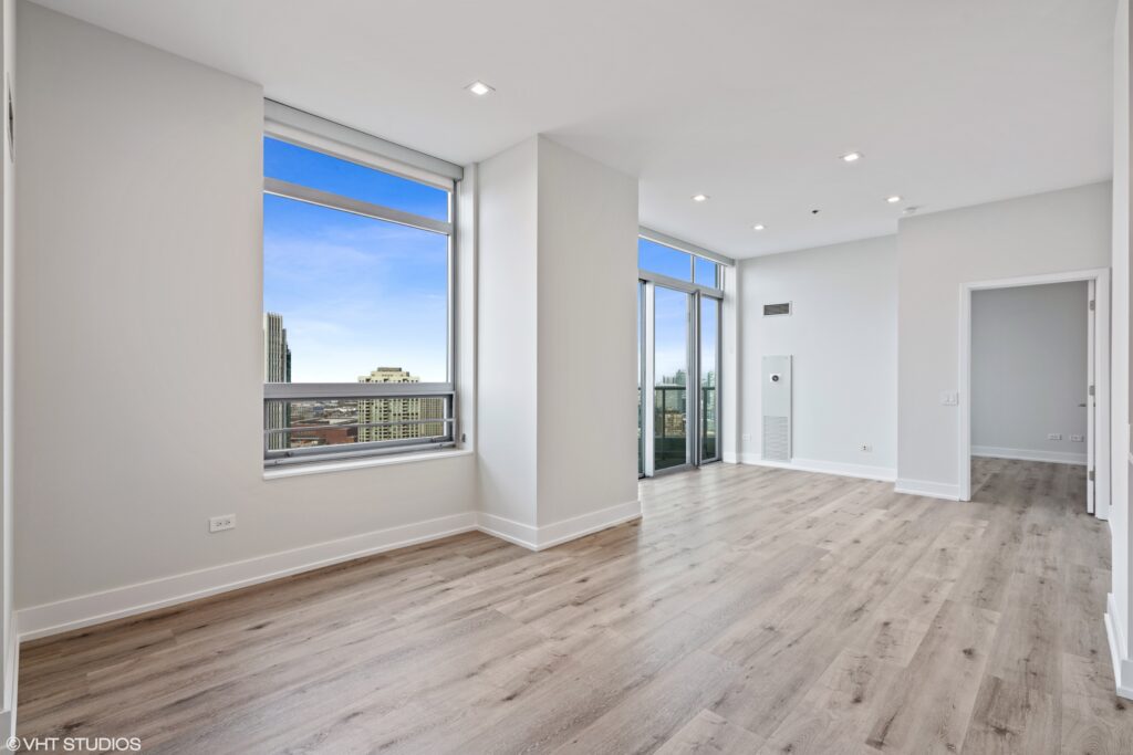Spacious, modern apartment interior with light wood flooring, large windows, and a sliding glass door leading to a balcony. Bright cityscape view under a clear blue sky.