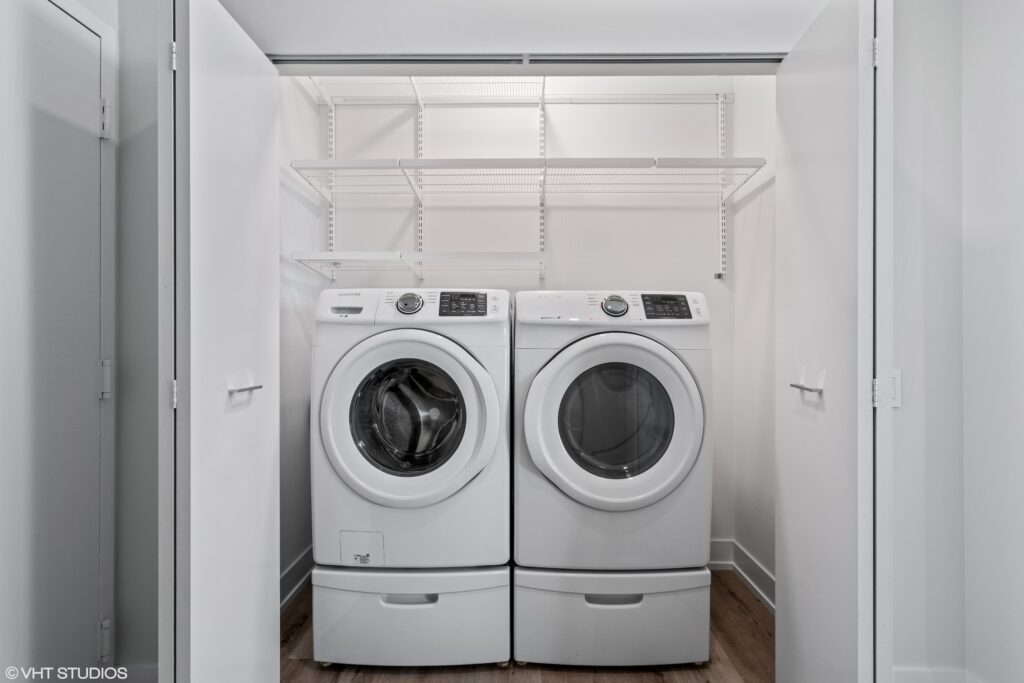 A laundry closet with a front-loading washer and dryer side by side, white shelves above, and double doors open to reveal the space.
