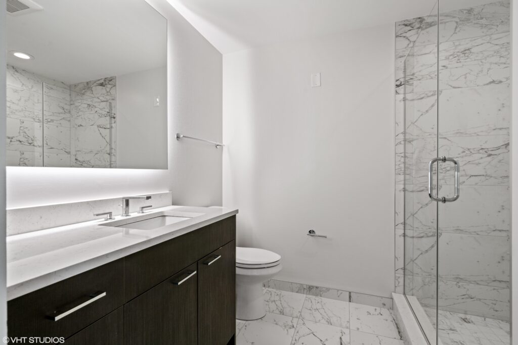 Modern bathroom with a marble shower, double sink vanity with dark cabinetry, lighted mirror, and a toilet.