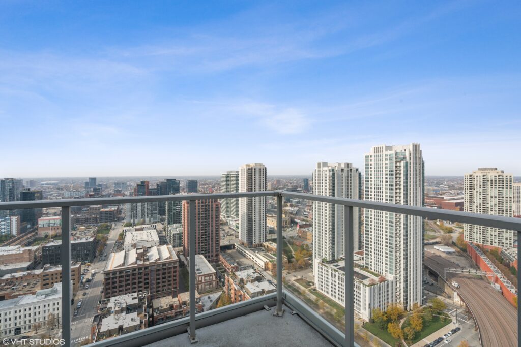 High-rise balcony view overlooking a cityscape with tall buildings, roads, and a partly cloudy sky in the background.