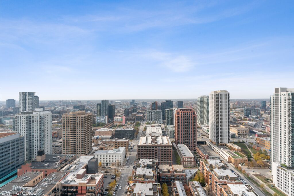 Aerial view of a cityscape with tall buildings, streets, and smaller structures under a blue sky with light clouds.