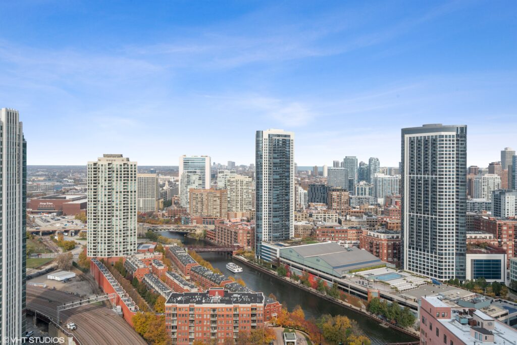 A cityscape view featuring tall modern buildings, a river with boats, residential complexes, and clear blue sky.