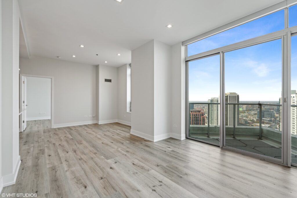 An empty modern apartment interior with light wood flooring, large windows, and a balcony overlooking a cityscape.