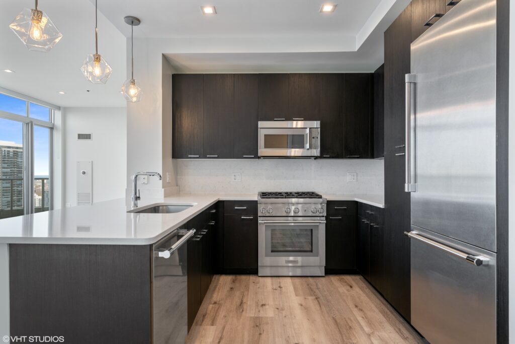 Modern kitchen with dark cabinets, stainless steel appliances, a large refrigerator, microwave, and oven. It features a white countertop with an island and three pendant lights overhead.
