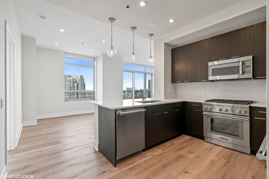 Modern kitchen with dark cabinets, stainless steel appliances, and an island, opening to a bright living area with large windows and a city view.