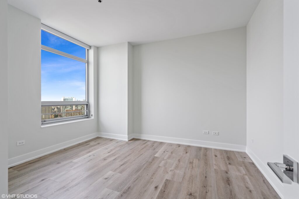 Empty room with light wood flooring, a large window showing a cityscape, and white walls.