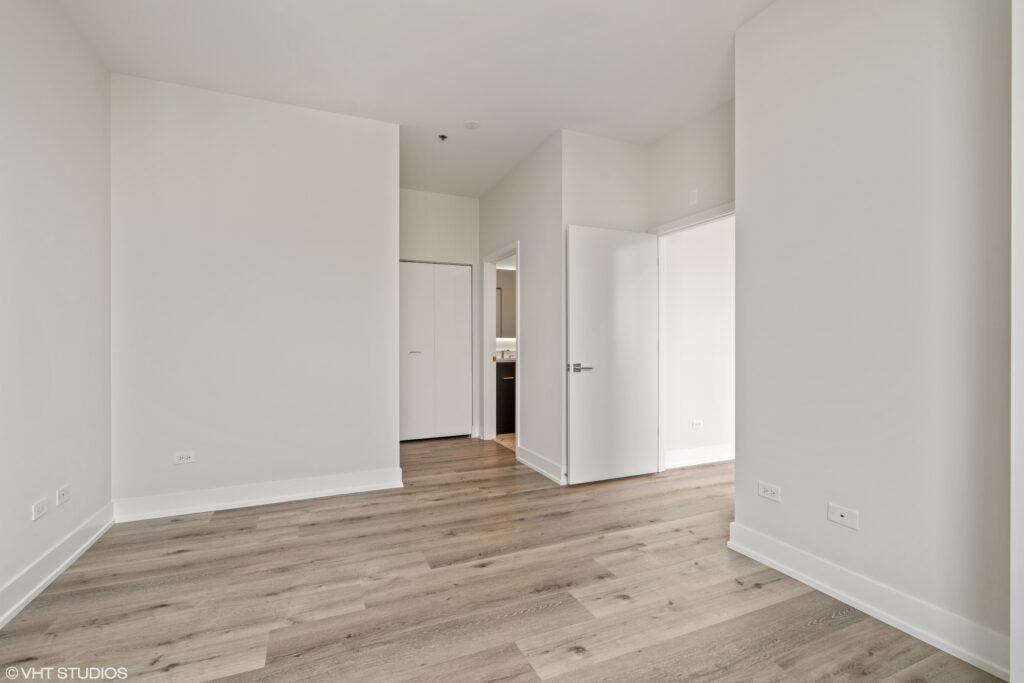 Empty room with light wood flooring, white walls, and two open doors. One door leads to a closet, and the other opens to a hallway with a glimpse of a kitchen.