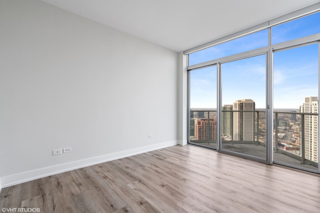 Unfurnished apartment room with light wood flooring, white walls, and floor-to-ceiling windows overlooking a city skyline with tall buildings.