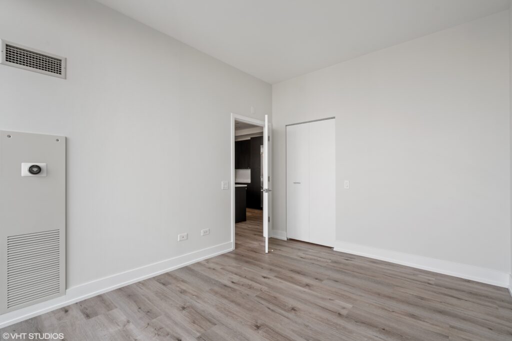 Empty room with light gray walls, wood-style flooring, an open door leading to a darker room, and a closed white closet door.