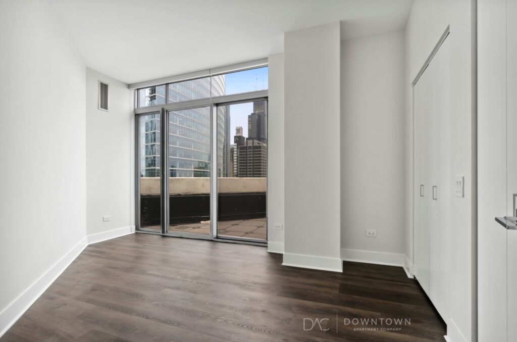 Empty room with large windows, wood floors, and a view of city skyscrapers. White walls and sliding closet doors are visible on the right.