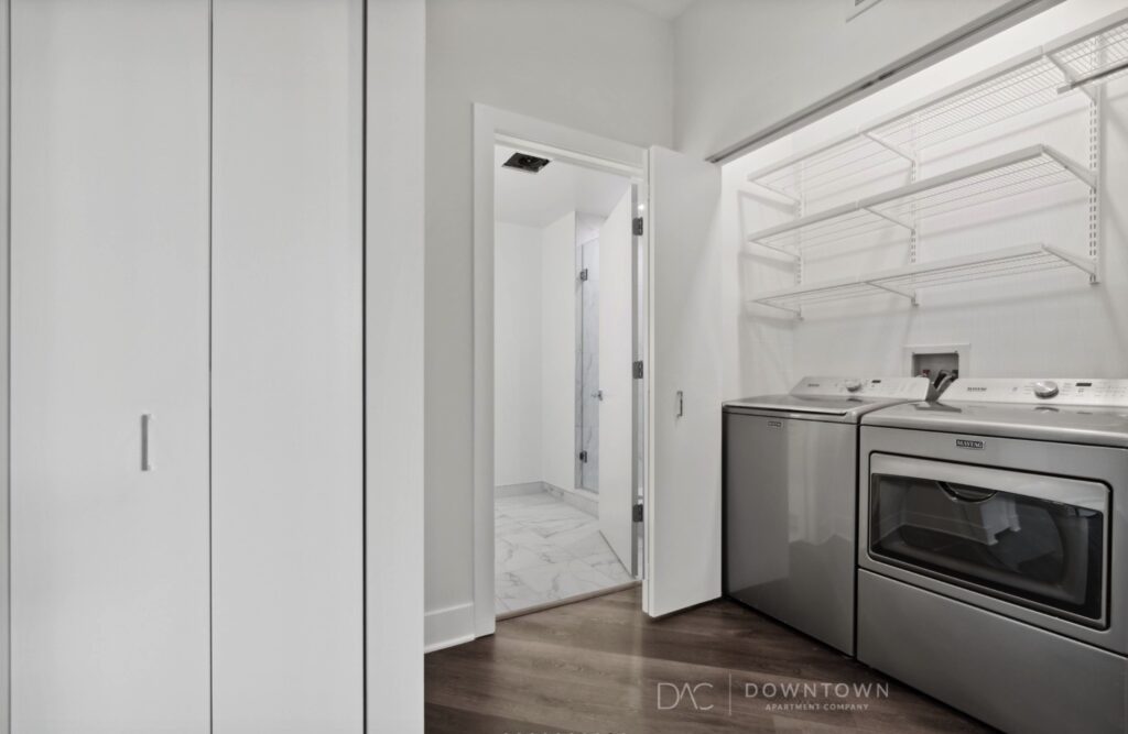 Laundry room with a washing machine and dryer on the right, open shelving above, and a door leading to a bathroom in the background.