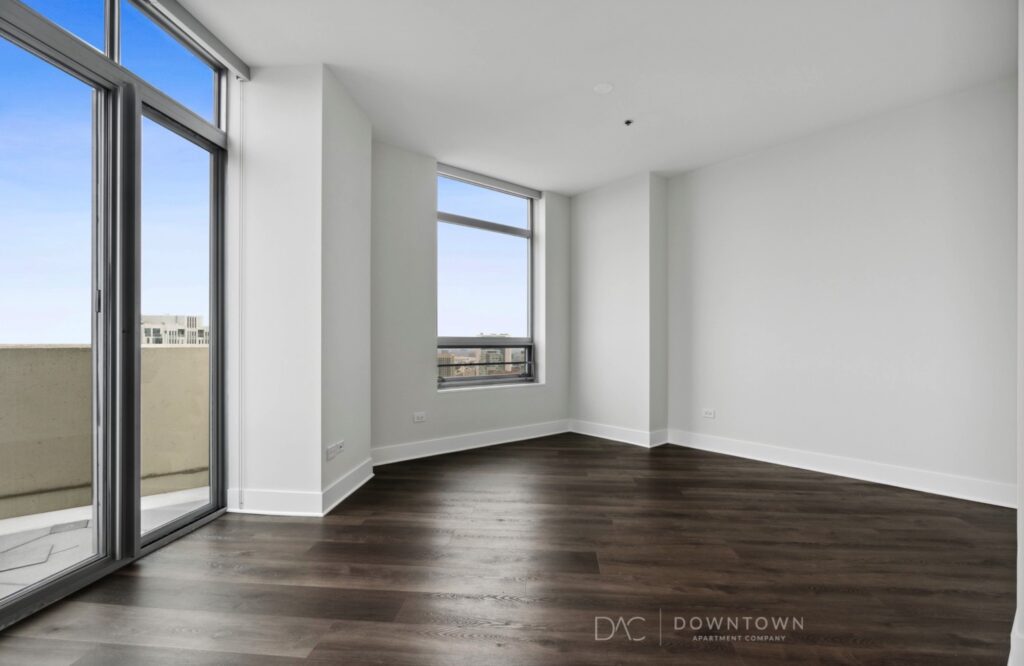Empty modern room with large windows, gray walls, and dark wood flooring, overlooking a cityscape.