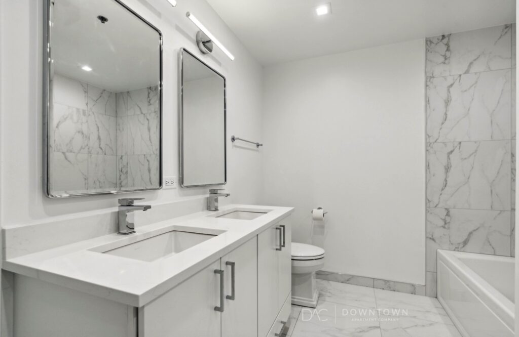Modern bathroom with two rectangular mirrors above dual sinks on a white counter. Marble tiles line the walls. A toilet and bathtub are in the background.