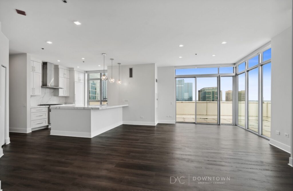 Spacious modern apartment with large windows, dark wood flooring, an open kitchen with white cabinets, and ceiling lights. Balcony offers a cityscape view.