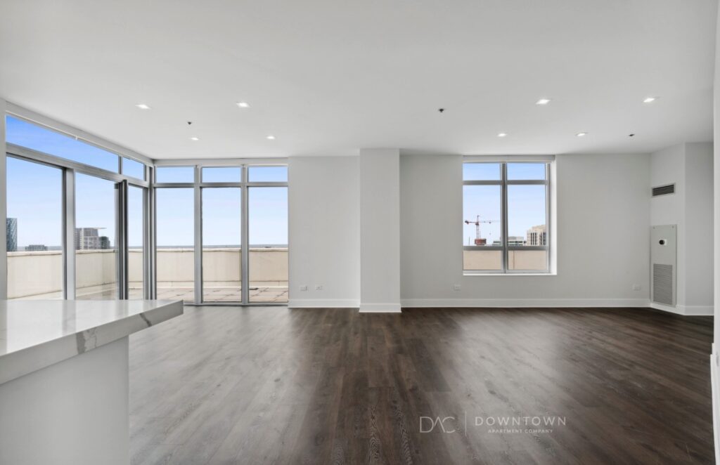 Spacious, empty apartment with large windows, wood floors, and a city view. White walls and recessed lighting.