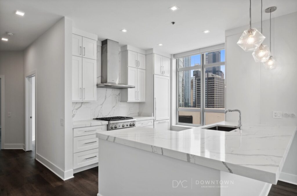 Modern kitchen with white cabinets, marble countertops, and stainless steel appliances. Large window shows city skyline. Three pendant lights hang over the bar area.
