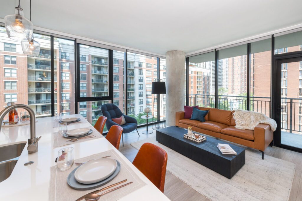 Modern open-concept living room with floor-to-ceiling windows, brown leather sofa, black coffee table, dining area, and city view.