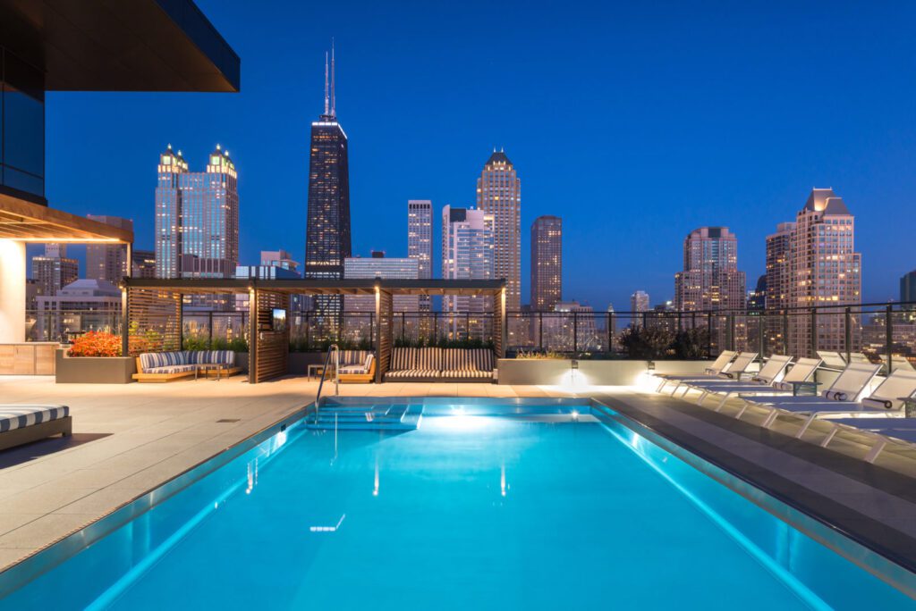 Rooftop pool at night with city skyline view, including tall buildings under a clear blue sky. Lounge chairs and shaded sitting area in the foreground.