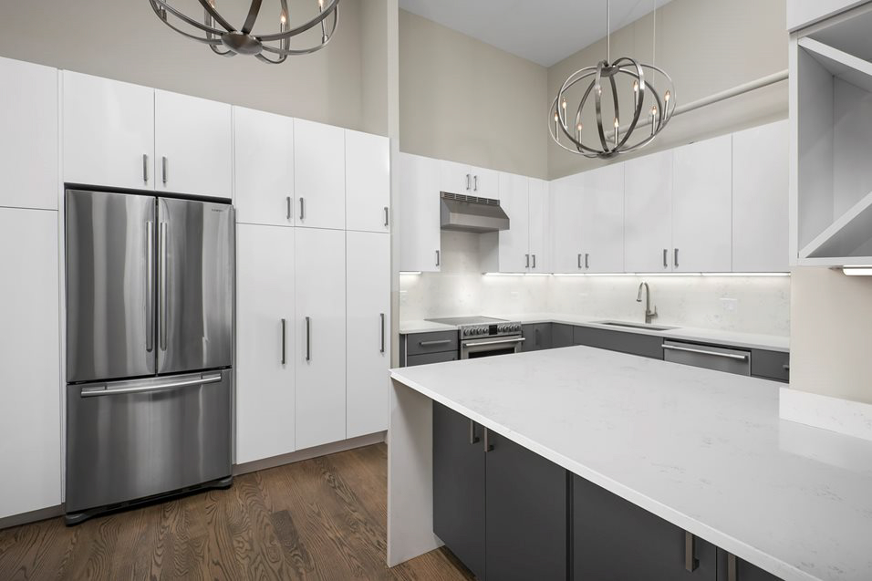 Modern kitchen with white and gray cabinets, stainless steel fridge, stove with hood, large island, hardwood floor, and spherical light fixtures.