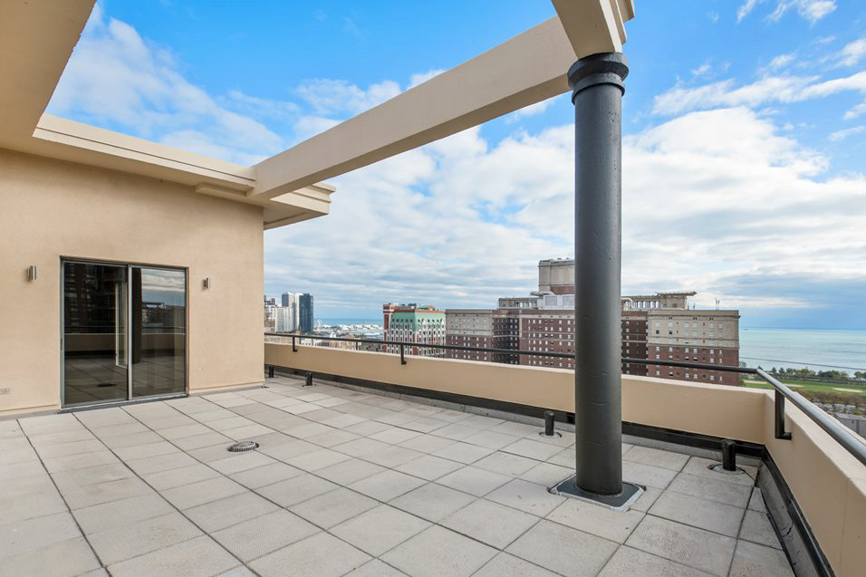 Rooftop terrace with a tiled floor, black support pillar, and glass door. Overlooks city buildings and a view of a large body of water under a partly cloudy sky.