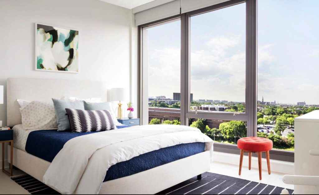 Modern bedroom with a large window showing a cityscape. A bed with blue and white bedding, a small red stool, and abstract art decorate the space.
