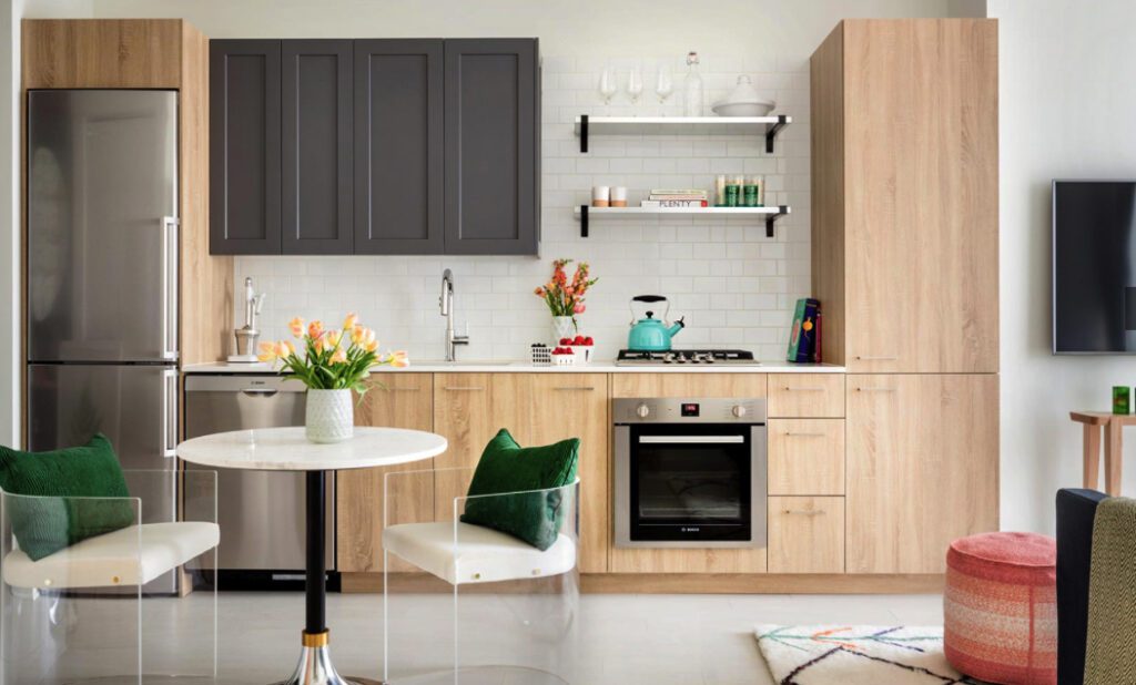 Modern kitchen with light wood cabinets, a stainless steel fridge, an oven, and a teal kettle on the stove. A round table with flowers and two transparent chairs is in the foreground.
