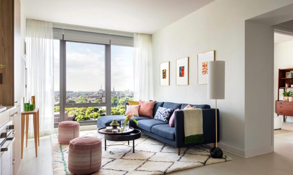 Modern living room with large window view, blue sofa, pink and green cushions, round coffee table, two pink ottomans, abstract wall art, standing lamp, and neutral-toned decor.