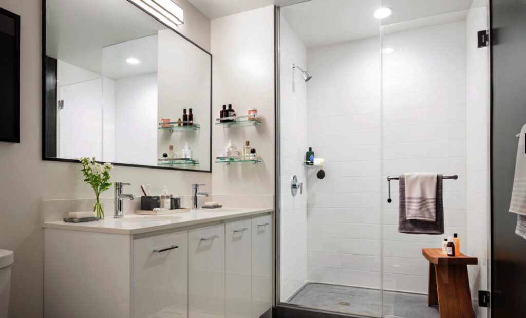 Modern bathroom with a glass-enclosed shower, white cabinets, wall-mounted shelves with toiletries, a large mirror, and a vase with flowers on the counter. Hanging towel and wooden stool visible.