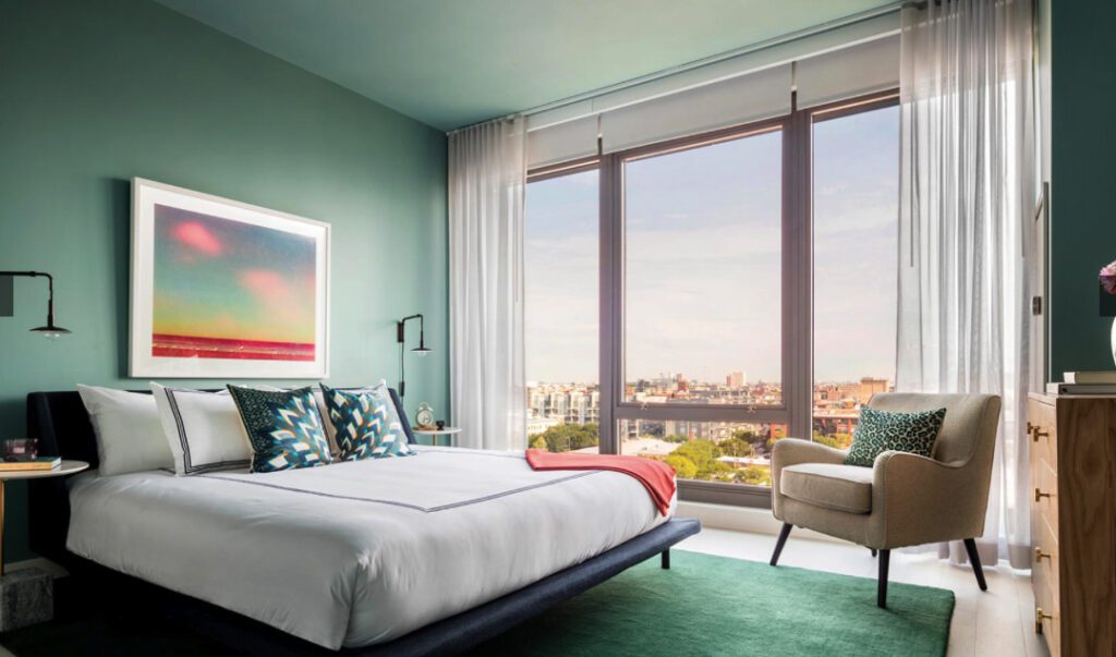 Modern bedroom with a large window view of a cityscape. Features a bed with colorful pillows, a framed artwork, an armchair, and a chest of drawers. Walls and ceiling are soft green.