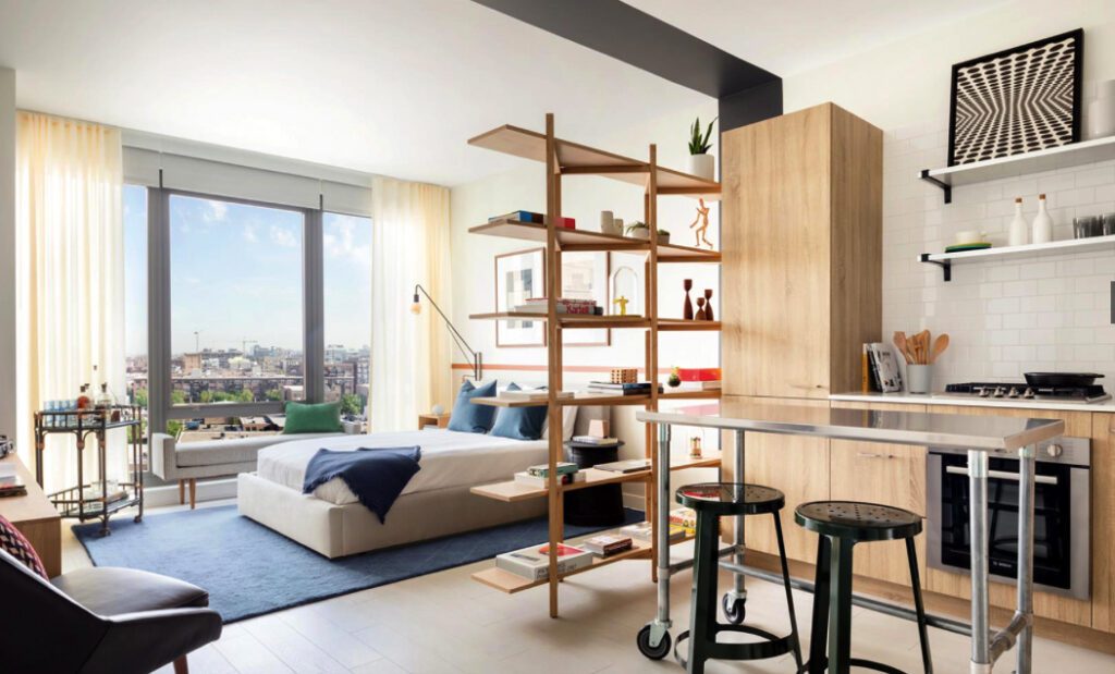 Modern studio apartment with a bed, dividing shelves, kitchen area, and large windows. Light wood furniture, white walls, and minimalistic decor create an open and airy atmosphere.
