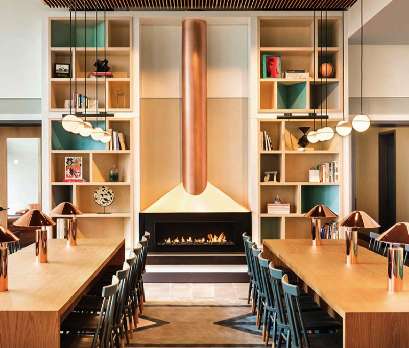 Modern dining area with wooden tables and chairs, a lit fireplace, pendant lights, and shelves with decorative items and books.