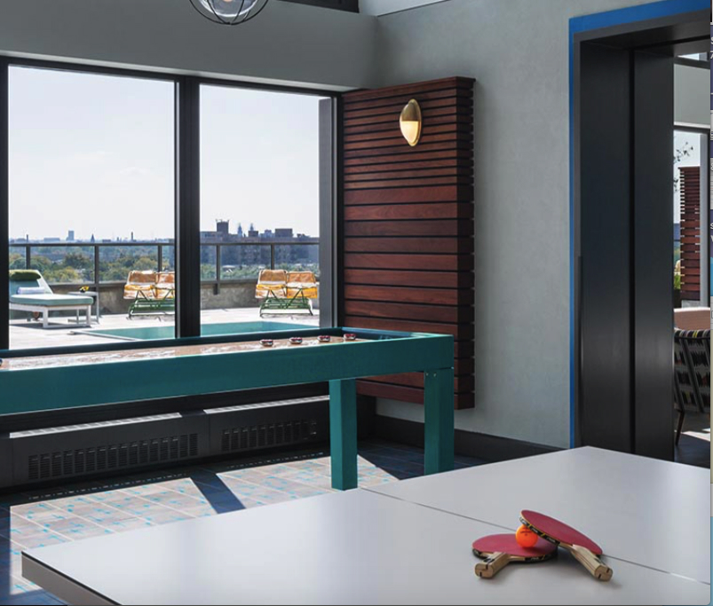 Room with a ping pong and shuffleboard table near large windows. Terrace with lounge chairs visible outside.