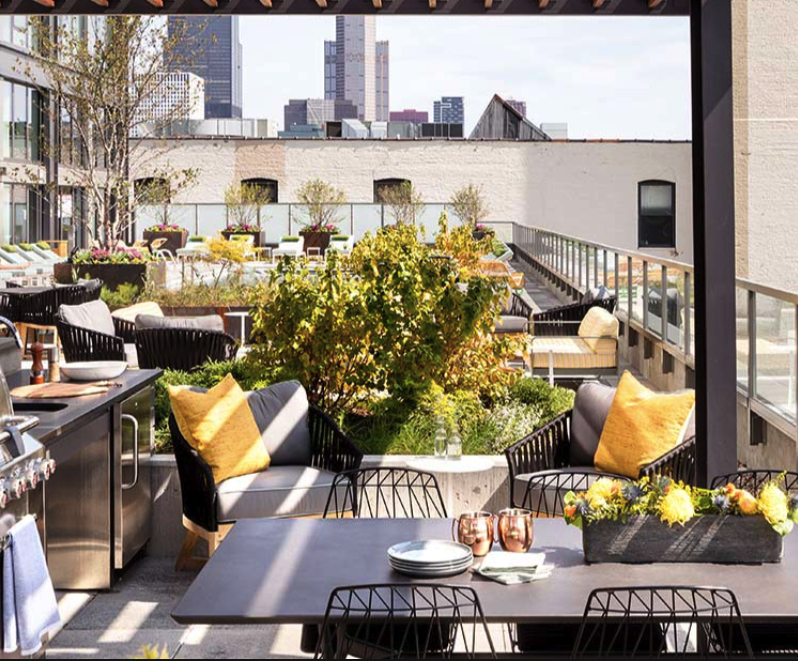 Rooftop patio with modern furniture, including tables, chairs, and sofas with yellow cushions. Urban skyline in the background. Lush plants and flowers decorate the space.