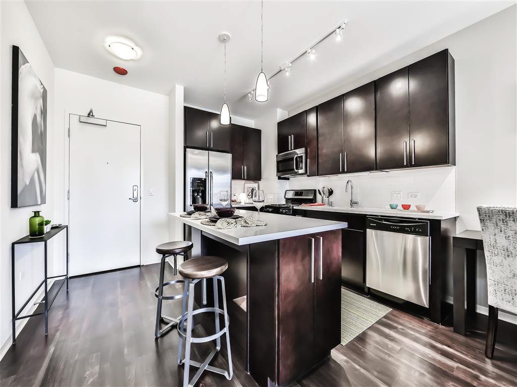Modern kitchen with dark wood cabinets, stainless steel appliances, and a large island with stools. White walls and a framed artwork on the left. Pendant lights hang over the island.