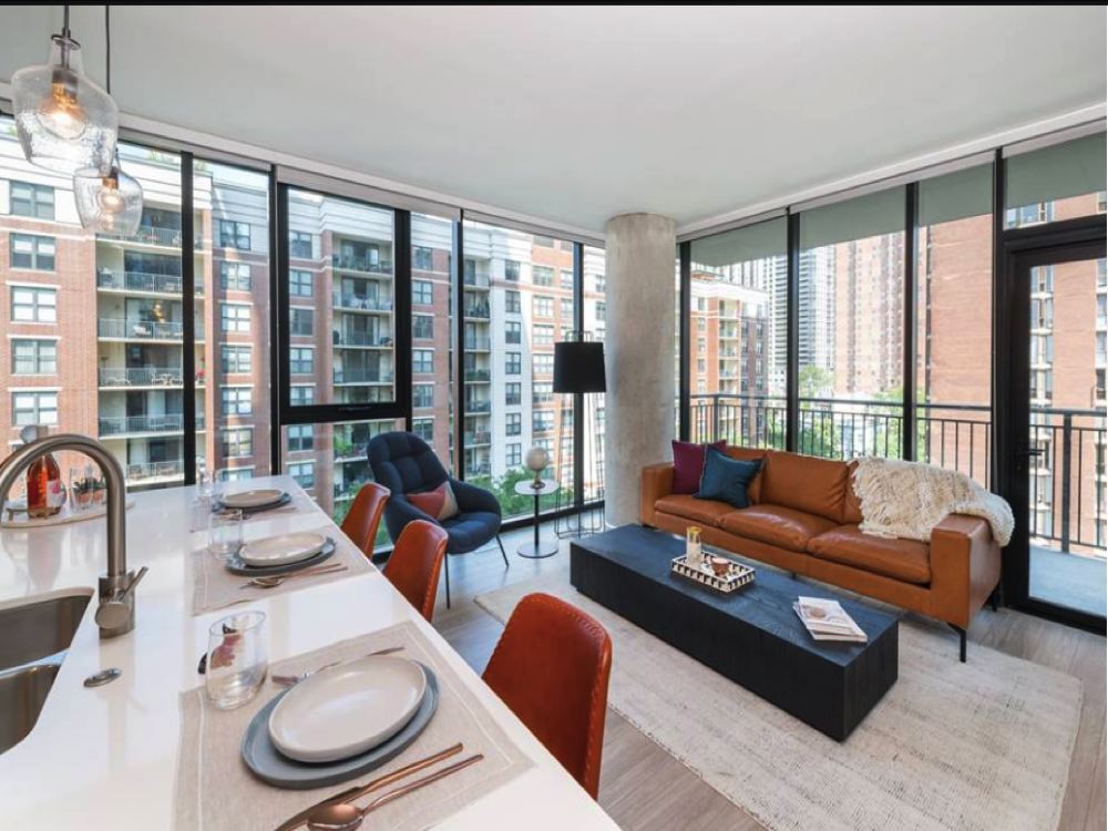 Modern apartment living room with large windows, brown leather sofa, black coffee table, and adjacent kitchen island. Urban view with high-rise buildings outside.