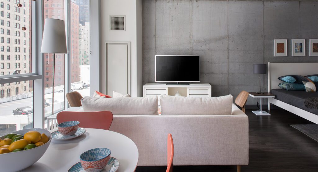 Modern studio apartment with a large window, white sofa, TV on a stand, round dining table with bowls, and a bed in the corner, featuring a concrete accent wall and city view.