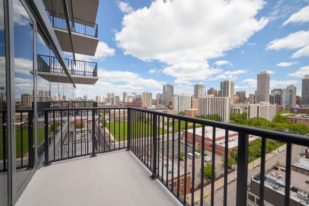 Cityscape view from a balcony showing buildings, a green field, and a partly cloudy sky.